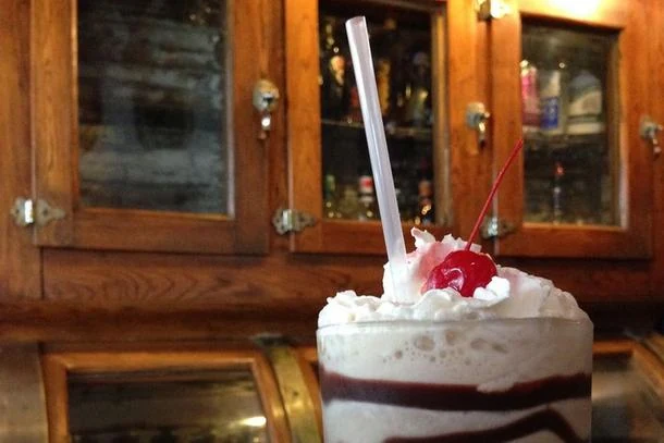 A Bushwhacker in a tall glass with whipped cream and a cherry, on the bar in front of glass-fronted oak back-bar cabinets