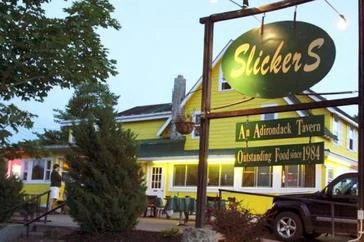 The lit oval Slickers sign at dusk in front of the yellow clapboard tavern on Route 28, reading "An Adirondack Tavern — Outstanding Food since 1984"