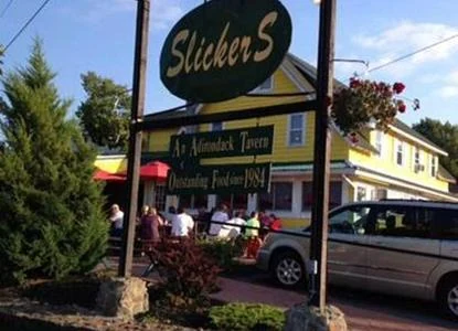 Guests at tables under red umbrellas on the deck in front of Slickers, with the oval sign overhead